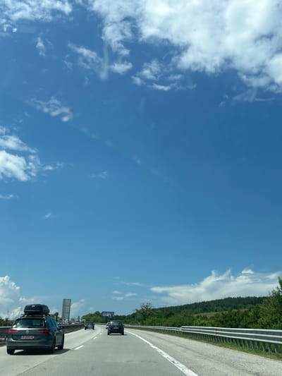 The image depicts a multi-lane highway scene on a bright, sunny day near the city of Ihtiman, Bulgaria, captured from the perspective of a vehicle moving forward. The sky occupies the upper two-thirds of the frame, exhibiting a vibrant blue hue with scattered white cumulus clouds. Several faint, translucent smudges or lens flares are visible in the sky area.

In the foreground, a dark grey Volkswagen Passat station wagon with a black Thule roof cargo box occupies the left lane of the highway. The vehicle's rear license plate, "CB 0045 HC", is clearly discernible. The asphalt road surface shows distinct white lane markings. Further ahead on the highway, multiple other vehicles are visible, including a dark sedan in the right lane and another dark vehicle in the left lane.

A silver metal guardrail runs along the right side of the highway, bordering a verdant landscape. The surrounding environment features dense green trees and vegetation, with a forested hill rising on the right side of the road and extending into the background. On the left side of the highway, a large, dark grey gantry structure is partially visible above the traffic. In the distance on the right, a large billboard with an advertisement is positioned above the tree line.