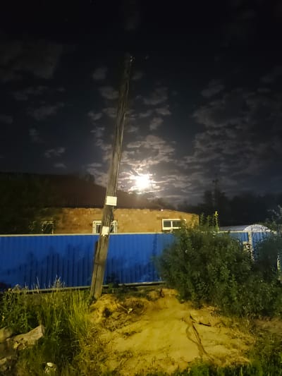 The image depicts a nighttime scene in Ekibastuz, Kazakhstan, illuminated by moonlight filtering through scattered clouds. A wooden utility pole stands prominently in the foreground, angled slightly towards the right. Two white rectangular signs are affixed to the pole. Behind the pole, a low, brick building with a reddish-brown, corrugated roof is visible. The building has multiple windows, some of which appear illuminated from within. A bright blue corrugated metal fence runs horizontally across the middle ground, separating the foreground from the background. To the right of the pole, dense green foliage and bushes obscure parts of the fence and the building. The ground in the foreground is a mix of dirt and dry grass, with some debris scattered around. The sky is dark, with soft, illuminated clouds suggesting the presence of a moon behind them. No people are visible in the image.