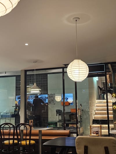 The image shows the interior of a restaurant in Ngaglik, Indonesia. The scene is illuminated by warm, artificial lighting. In the foreground, a table with chairs is set up, suggesting a dining area. Behind it, a glass partition reveals a glimpse into the kitchen, where a person in dark clothing is working. Several pendant lights hang from the ceiling, contributing to the ambiance. To the right, a set of stairs with integrated lighting ascends, hinting at a multi-level establishment. Various decorative elements, including paper lanterns and signage, are visible throughout the space, creating a cozy and inviting atmosphere.