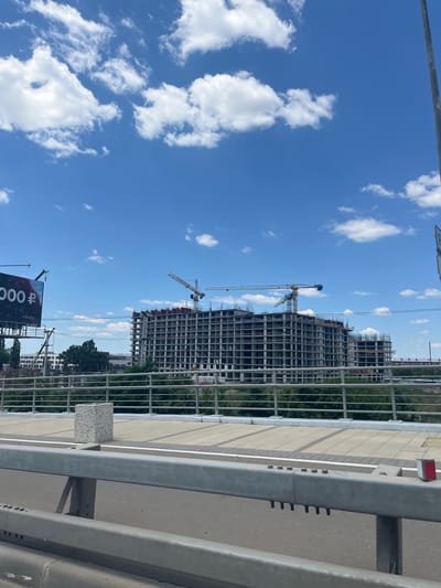 A construction site of a large apartment complex is visible under a bright blue sky with scattered white clouds. Two cranes are actively working on the multi-story building, which appears to be in the early stages of construction, with its concrete framework exposed. In the foreground, a pedestrian walkway with a metal railing runs along a bridge, with cars visible on the road below. A billboard with Russian text and a price is also visible on the left side of the image. The scene is set in Yablonovskiy, Russia, a city known for its rapid development and new construction projects.