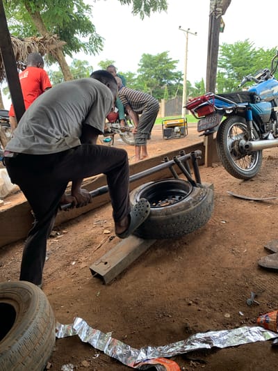 In Kabba, Nigeria, a man is using a metal bar to leverage a tire off a rim. He is wearing dark pants and a grey t-shirt, with one foot in a black sandal. The tire is on a stand made of metal and wood. In the background, two other men are visible. One is wearing a red shirt, and the other is wearing a striped shirt. A blue motorcycle is parked nearby, and a yellow generator is also present. The ground is dirt, and there are some scattered metallic wrappers. Trees and a utility pole are visible in the background. The scene appears to be an outdoor workshop or repair area.