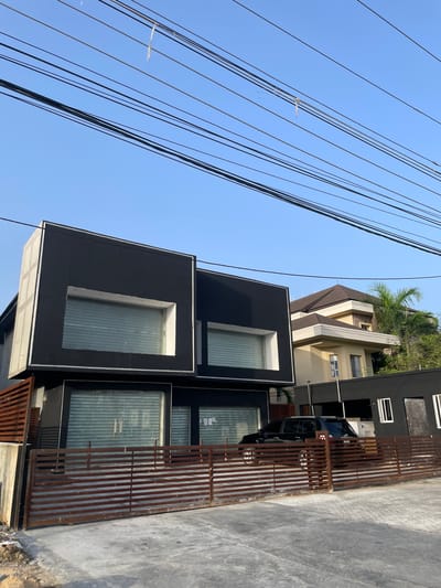 A low-angle, medium shot depicts a modern two-story building in Ilado, Nigeria. The building features a black facade and large, rectangular windows with security shutters. A rusted metal fence partially obscures a black SUV parked in front. To the left, a dense network of overhead power lines crisscrosses the clear blue sky. In the background, a portion of another building with a brown roof and beige walls is visible, topped with a palm tree. The foreground features a gray concrete surface. No people are visible.