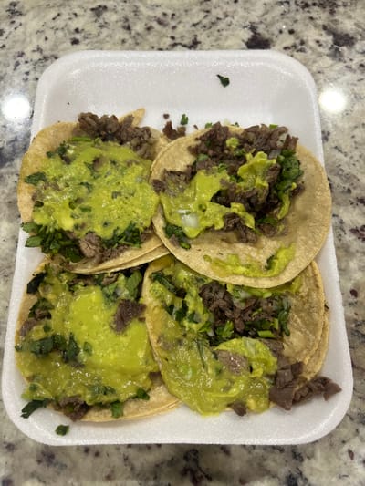 The image depicts four street tacos in a white, rectangular styrofoam container on a beige countertop with a speckled pattern. Each taco contains carne asada, cilantro, and what appears to be guacamole or avocado sauce. The tacos are arranged closely together within the container. The scene suggests a simple, informal setting, potentially a takeout meal. There is no explicit information to confirm the exact location as Barrio de Nuevo León, Mexico; this is an unsupported detail. No people are visible in the frame.