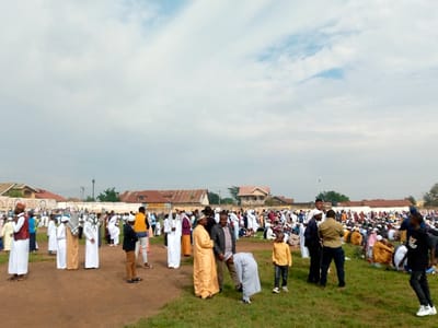 The image shows a large gathering of people in a field, likely in the city of Beni, Democratic Republic of Congo, at the coordinates provided. The majority of people are men, wearing long white or yellow robes, and many are wearing white caps. The field is covered in short, green grass and a few people sit on the ground in the distance. Behind the people are several buildings with red tile roofs, some are white and others are brown. The sky is cloudy, but the weather is mostly clear. The image appears to depict a religious gathering or celebration.