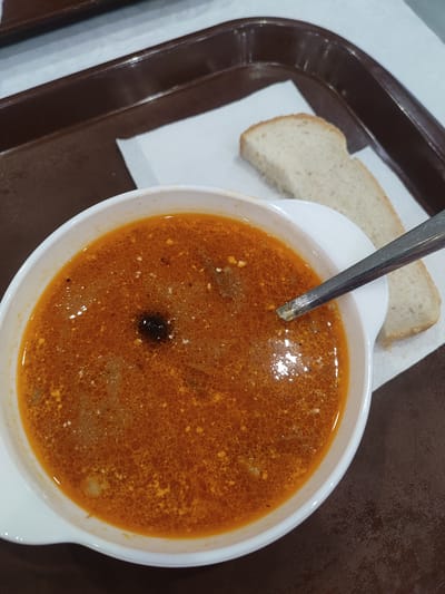A white bowl of vibrant orange soup with visible chunks of food and small white flecks of seasoning sits on a white paper napkin. A slice of bread rests to the right of the bowl, partially obscured by the soup bowl's rim. A metal spoon is placed in the soup, its handle extending out of frame. The bowl and napkin are on a dark brown rectangular tray, which is positioned on a light-colored surface, possibly a table. The overall scene suggests a meal, likely in a casual dining setting. The image was taken in Naberezhnye Chelny, Russia.