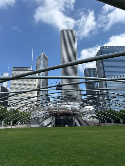 This image shows a view of Jay Pritzker Pavilion in Millennium Park, Chicago, Illinois, United States of America. The pavilion's distinctive stainless steel canopy structure dominates the foreground, with its parabolic arches curving over a large grassy lawn. In the background, several tall skyscrapers rise against a partly cloudy blue sky.
The pavilion itself features a stage area with a metallic, sculptural facade. Several speaker boxes are suspended from the canopy's framework. The lawn in front of the pavilion is lush and green, appearing well-maintained. A few people are visible in the distance on the lawn and near the pavilion, appearing to be casually walking. The overall scene depicts a public urban park space on a bright, clear day.