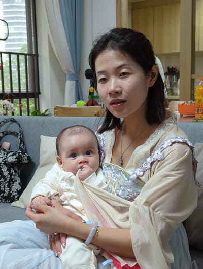 An indoor scene shows a woman holding a baby. The woman has dark hair and is wearing a light-colored top with embroidered detailing. She is looking at the camera. The baby is wearing white clothing and is partially covered by a light-colored cloth. The baby is looking at the camera. They are sitting on a grey sofa. In the background, there is a window with bars, part of a curtain, a shelf with bottles on it, a toy, and a black and white floral patterned bag. The location is potentially in the city of Shunde District, China, based on visual cues.