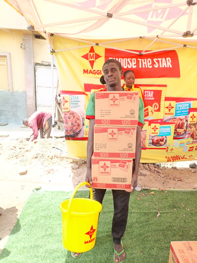 This media file captures an outdoor scene in Tarauni, Nigeria, likely during daylight hours given the bright lighting. The central subject is a man standing under a large yellow tent, holding a stack of Maggi seasoning boxes. He is looking directly at the camera with a neutral expression. Behind him, a woman is also partially visible, standing under the tent.

The tent itself is branded with "MAGGI" logos and the slogan "CHOOSE THE STAR". The background reveals a somewhat dusty and uneven ground, with a glimpse of a building on the left. Another person is seen in the background on the left, seemingly engaged in some manual labor, digging or clearing the ground.

The man is holding three boxes of Maggi Star Seasoning, with visible text indicating "20x100x4g", "Net weight 8kg", and production/expiry dates. He is also holding a bright yellow Maggi branded bucket in his left hand. The overall impression is of a promotional event or distribution taking place outdoors, possibly in a market or community area. The presence of the Maggi branding and products suggests a marketing or sales activity. The ground appears to be a mix of dirt and artificial turf, further indicating a temporary setup for an event.
