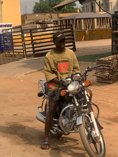 A dark-skinned male individual is seated on a black motorcycle on an unpaved, light brown road in Aguleri, Nigeria, under daylight conditions. He wears a black baseball cap with a gold Nike swoosh, an olive-green long-sleeved shirt with "HIGHVOGUE 27 FASHIONDEPT" text, dark ripped shorts, and black slide sandals. His hands are positioned on the motorcycle handlebars, and he gazes slightly to the right of the camera.

The motorcycle is black with numerous chrome accents, including the headlight, handlebars, engine guard, exhaust pipe, and a rear luggage rack. It features front and rear orange turn signals and a visible engine compartment.

In the background, to the left, is a yellow building partially obscured by a black and yellow striped metal gate. A blue sign on this building shows partial text, including "OINGS". To the right, a stack of what appears to be metal pipes or rods lies horizontally. Behind the gate and stack, multiple buildings in varying states of construction or completion are visible, including a light-colored structure with vertical wooden slats on its facade and a multi-story building further back. The sky is overcast or hazy.