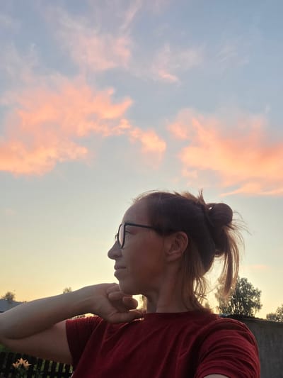 A woman with glasses and her hair in a bun, wearing a red shirt, looks to the left in profile against a sunset sky with pink clouds. The background shows trees and a fence. The scene appears to be in an outdoor setting in Буренка, Russia.