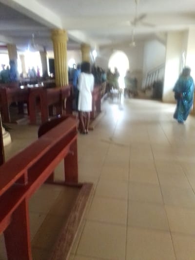 The image shows a large, empty room with rows of wooden pews along the walls. The room has a light-colored tile floor and a white ceiling. Several people are standing in the room, one of whom is wearing a white dress and is standing in the middle of the room. In the background, a person is walking away from the camera, wearing a blue dress and walking down a hallway. The image is taken from a low angle, giving the viewer a perspective from behind the pews in the foreground. The location of the image is a building in Auchi, Nigeria, based on the provided coordinates (latitude: 7.068, longitude: 6.269).