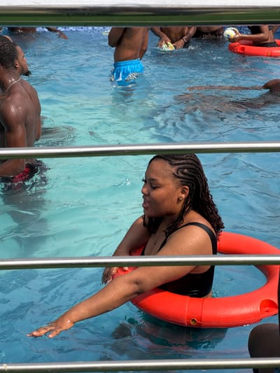 The image depicts an outdoor swimming pool scene in Eziobodo, Nigeria, under bright daylight. In the foreground, a young adult female with dark skin and braided hair is partially visible, positioned between two horizontal silver metal railings. She wears a black one-piece swimsuit, and an orange lifebuoy encircles her torso. Her right arm is extended, with her hand touching the light blue water surface. Her mouth is slightly open, and she looks towards her left. In the background, multiple individuals, predominantly males, are partially submerged in the pool. These include a shirtless male with a beard on the far left, another male wearing blue swim trunks in the middle, and several others holding various colorful buoyant objects, including a second orange float. All individuals are engaged in recreational activity within the pool.