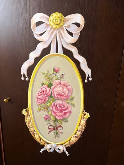 A painted floral arrangement featuring pink roses tied with a red ribbon is mounted on a dark brown wooden surface, possibly a door. The arrangement is framed by an oval, gold-colored border, adorned with decorative gold elements on the bottom. Above the oval frame, a white bow with a yellow centerpiece is depicted, with white ribbon tails extending downwards. Below the oval frame, another white bow is shown. The wooden surface has a visible wood grain texture and a small gold knob is visible on the left side. This object is located in Ozeretskoe, Russia.