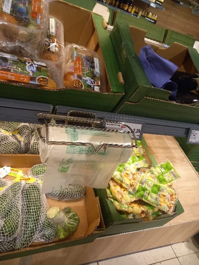 This image depicts a section of a supermarket or grocery store in Sallaumines, France. In the foreground, a metal dispenser holds a roll of clear plastic produce bags with green markings. Below it, to the left, a brown cardboard box contains several green avocados, most enclosed in white mesh nets. One avocado is unwrapped, positioned near a green, textured object resembling a broccoli crown wrapped in plastic. To the right of the dispenser, a green cardboard box holds numerous individually packaged, flat food items, possibly pastries or prepared meals, with green and white labels. These items rest on a light wood-grain display surface.

In the background, on a higher shelf, several products are displayed in open-top cardboard boxes with green exterior sides. To the left, multiple clear plastic-wrapped packages, likely containing produce such as potatoes ("pommes de terre" and "L'Or Hynois" visible on labels), are stacked. To the right, another green-sided box contains a folded blue fabric and several dark, elongated vegetables, consistent with eggplants. Further right, an additional green box is mostly empty. Price tags are affixed to the shelf structure, with one partially legible as "2,29." Distant shelves with various out-of-focus products line the background, indicative of a retail aisle. No people are visible.