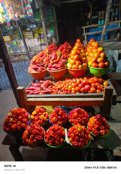A street vendor displays produce on a two-tiered wooden stand. The top tier holds peppers and tomatoes in colorful bowls, neatly arranged. The lower tier is filled with bowls of small, vibrant red and orange peppers. Behind the stand, a shop is stocked with goods on shelves. The scene is dimly lit, creating shadows around the edges of the frame.