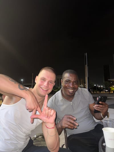 Two men are seated outdoors at night. The man on the left, with light skin and a buzz cut, wears a white tank top and has a tattoo on his left arm. He is smiling and making an "L" hand gesture with his right hand. The man on the right, with dark skin and short hair, wears a grey collared shirt. He is smiling and holding a black smartphone in his right hand. They are at a table with plastic cups and a white paper cup. In the background, city lights and buildings are visible, including tall, modern structures. The scene suggests an urban environment.
