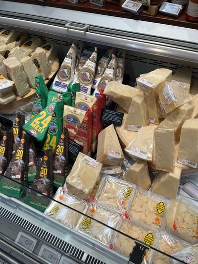 This image displays a refrigerated deli counter section in a grocery store in Monsummano Terme, Italy, dedicated primarily to various Italian cheeses. The brightly lit display case is filled with a dense assortment of cheese products, arranged on tiered shelves and behind a glass barrier.

The majority of the visible cheese consists of irregular, pale yellow blocks and wedges, typical of hard aged cheeses, individually wrapped in clear plastic film and bearing white adhesive price labels.

Behind these unwrapped portions, several types of pre-packaged cheeses are visible. These include:
*   Triangularly packaged portions of "PARMIGIANO REGGIANO BIOLOGICO" (Organic Parmigiano Reggiano) in green packaging, some marked "24 MESI" (24 months).
*   Triangularly packaged portions of "PARMIGIANO REGGIANO Bruna" in dark brown/black packaging, featuring an image of a calf and indicating "30 MESI" (30 months).
*   White, triangularly packaged portions labeled "MONTECCHIO MONTONE DELLA MUCCA BIANCA" (Montecchio Lamb from White Cow) with a cow head logo.
*   Red/burgundy packaged portions of "PARMIGIANO REGGIANO Delle Vacche Rosse" (Parmigiano Reggiano from Red Cows).

At the bottom of the display, multiple clear rectangular plastic containers are filled with pre-shredded or shaved cheese, each labeled "GRANA PADANO." A metal grate forms the base of the display, and digital price tags are visible on a rail beneath the products. The upper section of the display, partially visible, shows additional packaged items and price labels on a dark shelf. No individuals are present in the scene.
