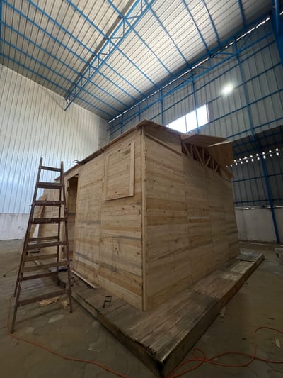 This media file depicts the interior of a large, unfinished structure, likely a warehouse or industrial building, in Gaza. The dominant feature is a tall, wooden construction resembling a two-story, multi-faceted room or booth, built from planks of light-colored wood. This structure appears to be in the process of being built or modified.

The setting is industrial, with a corrugated metal roof supported by blue metal beams, visible in the background. Natural light enters through a large opening in the roof, suggesting it's daytime. The floor is concrete and also appears to be a work in progress, with some areas covered in wooden planks. A rusty metal ladder leans against the wooden structure, indicating it's accessible from the ground level.

The wooden structure itself has a somewhat irregular shape, with angled walls and visible internal bracing made of wood, particularly on the upper section. A small, rectangular wooden panel is affixed to one of the walls, possibly a door or access hatch. An orange electrical cord snakes across the concrete floor near the wooden structure, suggesting the presence of power tools or lighting.

There is no visible text in the image. The overall impression is one of ongoing construction or renovation in a functional, albeit somewhat rough, environment. The scene is devoid of people, focusing entirely on the structure and its surroundings. The time of day is clearly daytime due to the natural light. The location cues, such as the style of the building and the context provided, point to Gaza, Palestinian Territories. There are no strong emotional cues, as the image focuses on the physical aspects of the construction.