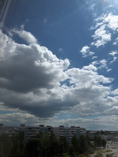 The media file depicts a sky filled with a variety of clouds, ranging from bright, puffy white cumulus clouds to darker, more ominous storm clouds. A thin wire stretches diagonally across the top left of the frame, suggesting the presence of power lines.
Below the sky, a residential area is visible, characterized by multi-story apartment buildings and mature trees. The architecture of the buildings, with their numerous balconies and windows, is typical of Soviet-era urban planning. The presence of green spaces, including what appears to be a park or lawn in the foreground, indicates a relatively well-developed urban environment. A street with parked cars can be seen further in the distance.
The lighting suggests it is daytime, likely late afternoon or early evening, given the mix of bright sunlight and shadowed clouds. The weather appears to be changeable, with both sunny and cloudy conditions present simultaneously. The scene is set in an urban environment, likely a residential district. There is no visible text in the media file. The overall mood is one of a typical day in a city, with the dynamic sky adding a sense of drama.