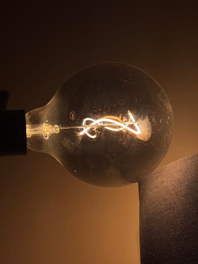 A dimly lit, close-up shot features a vintage-style light bulb with a visible filament, possibly located in Santa Clara Ocoyucan, Mexico. The bulb's glass is slightly dusty, and it's mounted on a black base. A portion of a lamp shade is visible on the right side of the bulb. The background is a muted brown color, suggesting an interior setting. No people or interactions are visible in the image.