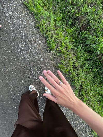 The image shows a person's legs and hand from a first-person perspective. The person is standing on a gravel path adjacent to a grassy area. The person is wearing dark brown trousers and white Crocs with cartoon characters. The person's right hand is extended towards the camera, revealing two rings on the ring finger. The background consists of lush green grass, suggesting an outdoor, possibly rural or park-like setting. The overall lighting indicates it is daytime, likely late afternoon or early evening due to the soft light. There is no specific indication of the location being within the city of Podshivalovo, Russia, as the image lacks any identifiable landmarks or context clues specific to that location.