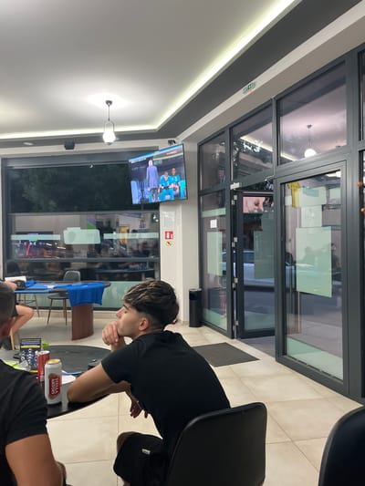 A group of young men are seated at a table in a modern cafe or bar in Kyustendil, Bulgaria, watching a television screen that is displaying a sports broadcast. The cafe has large glass windows and doors, with a sleek, contemporary design. The lighting is soft, with recessed ceiling lights and a prominent pendant lamp. One man in the foreground is wearing a black t-shirt and shorts, his hand resting on his chin as he gazes towards the TV. A Stella Artois beer can and a red can are on the table. Other patrons are visible in the background, also engaged with the television. The overall atmosphere appears relaxed and casual.