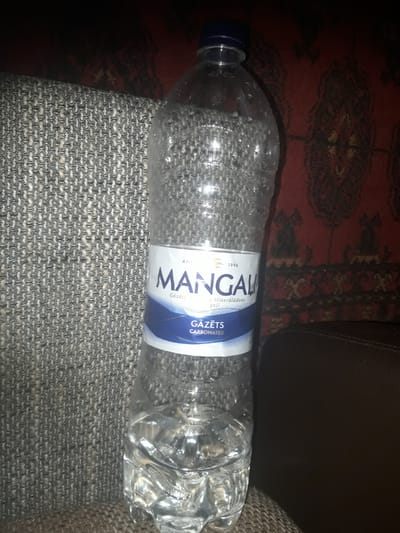 A plastic bottle of Mangali carbonated mineral water is displayed prominently against a textured, dark background. The bottle is partially filled with clear liquid and condensation is visible on its surface. A blue cap seals the bottle. The label on the bottle is white with blue accents and text that reads "Mangali" and "Gāzēts Carbonated". The background features a patterned dark fabric and a dark leather surface. The scene appears to be indoors, likely in a residential setting in Zilupe, Latvia, as indicated by the brand of water. There are no people or discernible actions captured in the image.