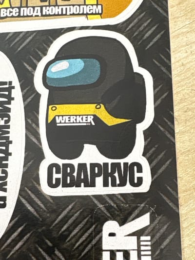 This media file features a close-up view of a sticker adorned with a cartoonish astronaut-like figure. The figure is primarily black and yellow, with a light blue visor. Text in Russian is visible on the sticker: "CBAPKYC" is written vertically on the right side of the figure, and the word "WERKER" is printed horizontally on a yellow section of the figure. Another sticker with Russian text, "все под контролем" (meaning "everything under control"), is partially visible on the left. The background of the sticker has a metallic, textured pattern resembling diamond plate.
To the right of the main sticker, part of another sticker is visible with large, white letters that appear to spell "R E R". Beneath the main sticker, another line of text is partially visible, seemingly in a stylized font. The overall aesthetic suggests these are decorative stickers, possibly applied to a surface like a laptop, notebook, or tool case.
The background of the image is a light-colored, textured surface, possibly a table. The lighting is even, and there are no strong shadows, indicating it was likely taken indoors or in diffused light. The image is cropped tightly on the stickers, providing no information about the larger environment or any people present. The sticker's design might allude to gaming or pop culture references, given the astronaut-like character and bold lettering. The presence of Russian text and the provided location context of Mahilyow, Belarus, further supports this.