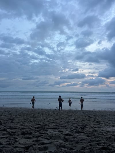 Four men are playing soccer on a sandy beach at sunset. The men are silhouetted against the cloudy sky. The ocean is in the background, and there are waves crashing on the shore. The scene is located on the beach near Playa Naranjo in Santa Fe, Costa Rica (Latitude: 9.642, Longitude: -85.168).