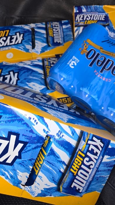 The image features boxes of Keystone Light beer with a blue and white wavy design and a yellow stripe. The brand name is prominently displayed. A blue 3-pack of Modelo beer cans rests on top of the Keystone Light boxes. Some text includes "ALWAYS SIG" and "CERVEZA". The close-up shot highlights the packaging details and color schemes of both beer brands.