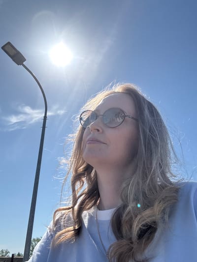 A woman wearing sunglasses and a light blue top is photographed from a low angle against a bright blue sky with the sun visible. A street lamp is also in the frame. The location cannot be determined as Balezino, Russia from the provided image.