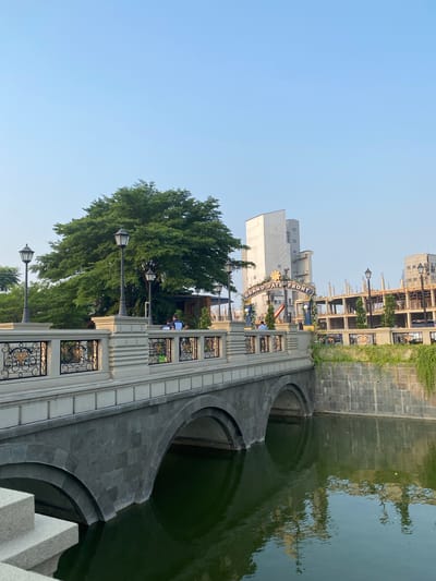 The image depicts the Grand Water Front in East Jakarta, Indonesia, an area characterized by a picturesque bridge spanning a body of water. The bridge, constructed with dark stone arches and a stone balustrade adorned with decorative metalwork, stands as a prominent feature. Lampposts line the walkway of the bridge, casting a warm glow. Beyond the bridge, the landscape reveals a large, leafy tree providing shade, and several buildings, some of which appear to be under construction. A prominent sign archway reads "GRAND WATER FRONT". A few individuals are visible on the bridge and near the buildings, suggesting a public or commercial space. The water beneath the bridge reflects the surrounding structures and greenery, adding to the serene ambiance of the scene. The sky is clear and blue, indicating a pleasant day.