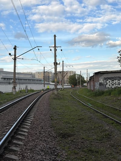 The image depicts a railway track in Lyubertsy, Russia, on a cloudy day. The perspective is from ground level, looking along the tracks as they curve to the right. The sky is partly cloudy, with patches of blue visible. In the foreground, the railway track is made of gravel and concrete sleepers.
To the left of the tracks, there are several utility poles with electrical wires strung between them. In the background, there are multi-story apartment buildings and a tall industrial chimney. There are also some hints of urban decay, with a brick building covered in graffiti.
There appear to be a couple of individuals in the distance, one wearing a yellow vest, possibly working on or near the tracks. The overall impression is one of an industrial or semi-urban environment on a day with mixed weather conditions. The graffiti on the brick building is a notable detail, featuring stylized white lettering. The time of day appears to be late afternoon or early evening, given the angle of the light.