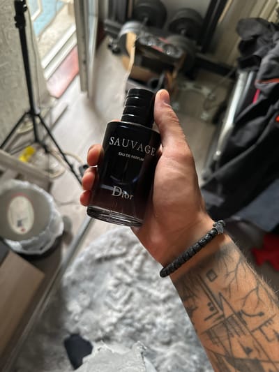 The image shows a close-up of a hand holding a bottle of Dior Sauvage Eau de Parfum. The bottle is dark glass with white lettering. The hand has several tattoos on the forearm and is wearing a black beaded bracelet. In the background, partially visible, are gym equipment, clothing, and a tripod with a light. The floor appears to be light-colored wood. The scene does not provide any information about the city of Kazanlak, Bulgaria.