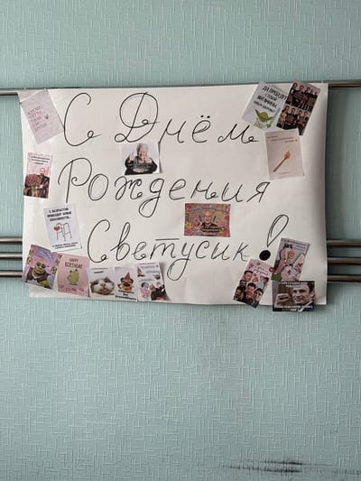 The image shows a handmade birthday poster affixed to a light blue, textured wallpaper. The poster is decorated with handwritten text and various printed images. The central handwritten text reads "С Днём Рождения Светусик!" which translates to "Happy Birthday Svetusik!". Around this central message, numerous smaller printed images are attached. These include greetings, humorous pictures, and photos of people. The poster is positioned horizontally, slightly above the midpoint of the visible wall, and appears to be held up by a metal rod. The wallpaper has a repeating geometric pattern. There is no indication of Mahilyow, Belarus, or any specific city in the image.