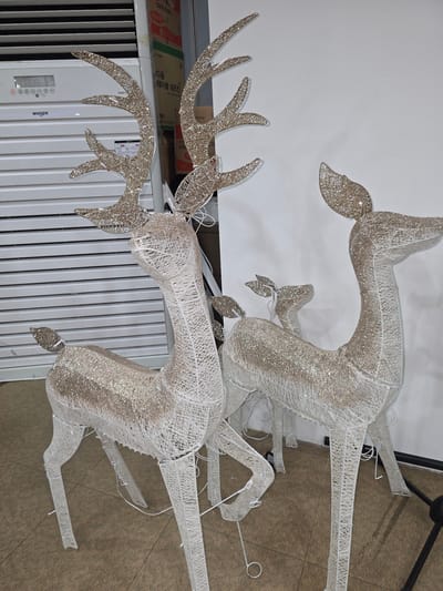 This image depicts a display of three wireframe reindeer decorations, two large and one small, covered in shimmering material, standing on a textured carpeted floor in front of a white wall and a white air conditioning unit. The larger reindeer on the left is positioned in the foreground, facing the viewer. Its body and legs are constructed from white wire mesh, and its antlers and back are adorned with glitter. To the right and slightly behind it is another large reindeer, also made of wire mesh and glitter, facing to the right. A smaller reindeer is visible between the two larger ones, nestled behind the one on the left. The air conditioning unit in the background has horizontal slats and a digital display showing "88°C". Cardboard boxes are stacked to the right of the air conditioner. This scene appears to be located indoors, likely in a commercial or residential setting, possibly Asan-si, South Korea, given the context.
