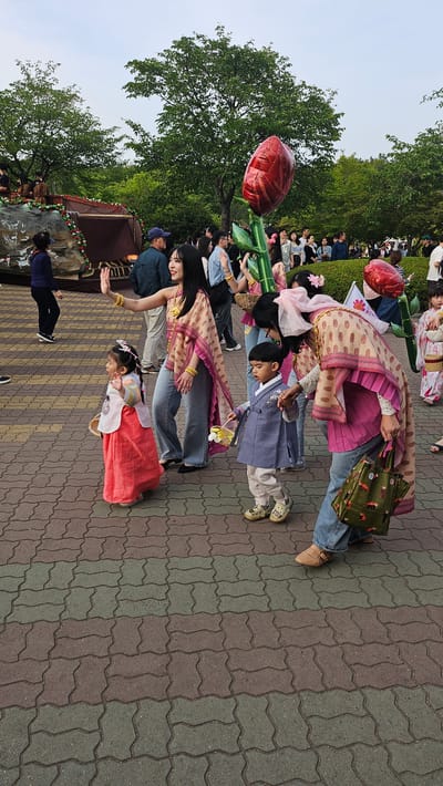 A medium, eye-level shot, rotated 90 degrees clockwise, depicts a group of people posing for a picture on a paved area in the city of Ulsan, South Korea. The pavement is composed of grey, brown and tan interlocking brick tiles. In the foreground, a woman wearing a beige and pink patterned top with blue jeans and gold bracelets poses next to a girl in a red and white traditional Korean hanbok. Behind them, another woman in a similar patterned top and jeans poses next to a boy in a light blue hanbok. An older woman in similar patterned top and jeans poses further back. They are holding small, ornate baskets. Behind them is a crowd of people observing. A large red rose balloon is held by someone in the crowd. To the left of the frame, a small decorative waterfall feature is visible, with a person in dark clothing standing near it. Trees and green foliage are visible in the background. The sky is overcast.