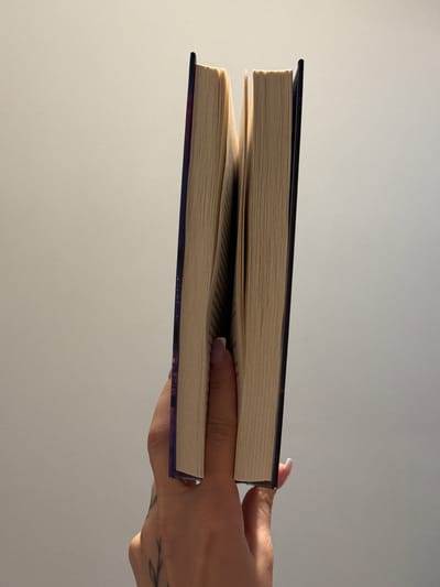 A person's hand is holding two books, one stacked on top of the other. The books are open, and the pages are visible, showing text. The top book has a dark cover, while the bottom book has a purple cover with some colorful patterns. The hand has fingernails with light pink polish and a tattoo of a plant on the wrist. The background is a plain, light gray wall, suggesting an indoor setting. The lighting appears to be soft, possibly from natural light coming from a window, as indicated by the subtle shadows. The image is taken from a slightly elevated angle, looking down at the books and hand. No specific time of day or weather conditions can be inferred. The location context of Łódź, Poland, does not provide any specific visual cues within the image itself.