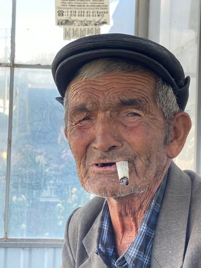 The image is a close-up portrait of an elderly man wearing a dark cap, a grey jacket, and a blue plaid shirt. He is smoking a cigarette, which is held in his mouth and lit. The man has weathered skin with deep wrinkles, grey hair, and a beard. He is positioned in front of a window with a pale blue tint, through which blurred foliage and some white flowers are visible. A piece of paper with Cyrillic text and a phone number is attached to the upper right side of the window. The overall lighting suggests an outdoor setting during the day. The location is stated to be Krasnomakskoye rural settlement, Ukraine.