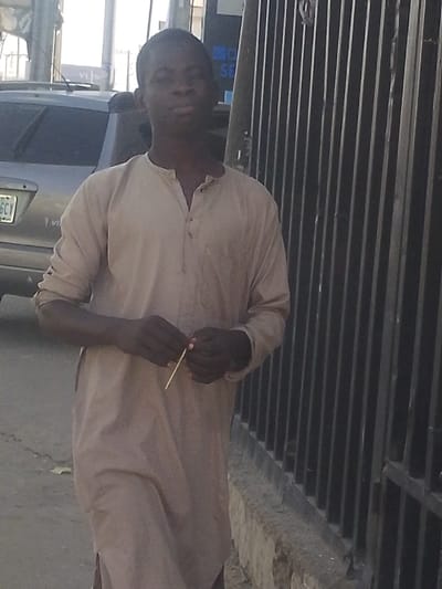A male individual with dark skin and short dark hair stands facing forward in a daytime outdoor setting in Babban Giji, Nigeria. He wears a light brown, long-sleeved tunic with a v-neck and three visible buttons. Both hands hold a thin, light-colored stick horizontally at waist level. To the subject's left, the rear portion of a silver or light-colored SUV is parked, displaying a Nigerian license plate with "66CY". To the subject's right, a dark metal fence composed of vertical bars extends vertically. The ground beneath the subject appears to be a concrete sidewalk or paved surface.
