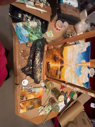 The image provides an overhead view of a busy artist's workbench, centered around a painting on an easel. The painting depicts a striking sunset or sunrise over water, rendered with thick, textured brushstrokes, particularly visible in the bright white clouds and golden light rays piercing through. Three boats, two motorboats and one sailboat, are silhouetted on the shimmering golden water, with distant land visible on the horizon.
The wooden workspace is covered with various art supplies and household items. To the left, a black tray holds numerous paint tubes and a palette knife. A small, abstract painting with green and blue impasto strokes lies nearby. Other items in this area include a roll of brown tape, a clear plastic bag with packing material, a white bag with "ALDI" branding, and a wooden board.
To the right of the easel, there are multiple glass jars, some used for paint or brushes, one with a "MAILLE" label partially visible. A green tape roll, a craft knife, red-handled scissors, and an "amazon" branded cardboard box are also present, along with a large roll of brown paper. Below the easel, a paint-stained black cloth is draped over the table. Two small boards covered in dried, colorful paint serve as palettes. A partially visible white tube is labeled "OIL PAINT".
The photograph is taken from a high angle, looking down onto the workspace. The artist's feet and the lower part of a pink garment are visible at the very bottom edge, suggesting the artist took the photo themselves while standing over their work. The scene conveys a lived-in, creative environment, actively used for painting.