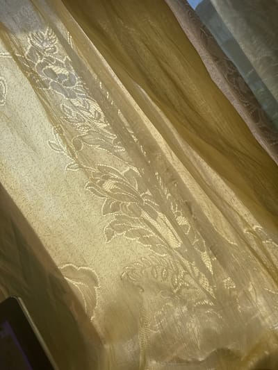 The image shows a close-up of sheer, golden curtains with a floral lace pattern. The sunlight is filtering through the fabric, creating shadows and highlighting the texture of the material. The curtains are draped, with visible folds and pleats, suggesting they are hanging in a window. In the lower left corner of the image, a dark object, possibly a tablet or e-reader, is partially visible. The scene is likely indoors, in a domestic setting, in the city of Bezden, Bulgaria.