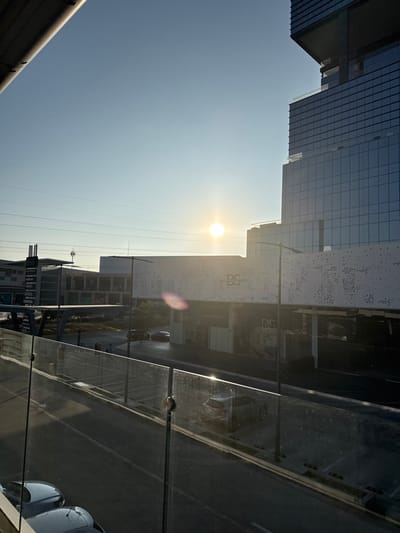 The image depicts an urban landscape under a clear, pale blue sky with a low, intensely bright sun positioned slightly right of center, casting strong light and a lens flare. The scene is viewed from an elevated position behind a clear glass balustrade with metal fasteners. An angular, grey structural element partially obscures the upper left corner.
In the mid-ground, a broad asphalt surface, marked with white lines indicating parking spaces or lanes, stretches across the scene. Several vehicles are present: parts of two silver/grey cars are visible in the bottom-left foreground, and a dark grey SUV is positioned in the mid-ground asphalt area, reflecting sunlight. Another dark vehicle is visible further back. A tall, black street light pole stands in the mid-ground, slightly right of center.
Dominating the right side of the frame is a modern, multi-story glass skyscraper with a dark framework, reflecting the sky. Adjacent to it, in the center-right, is a large commercial building featuring a light-colored facade with an irregular pattern of circular perforations. The letters "DS" and "BS" are visible on this perforated facade. Below this, darkened storefronts are present. To the left of this central building, an elevated, grey, angular structure, possibly a pedestrian bridge or ramp, is visible, partially obscuring additional commercial buildings in the background. Partial text, resembling "DOMTELAS," is discernible on one of these background buildings. Several indistinct human figures are visible on the ground level, near the commercial buildings.
Based on the architectural features, including the perforated facade, modern glass towers, and visible branding elements, the location is identified as the **Angelópolis Lifestyle Center area in Puebla City, Mexico**.