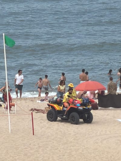 This image depicts a sunny day at a beach, likely in Espinho, Portugal, given the presence of beachgoers and the general coastal environment. In the foreground, an all-terrain vehicle (ATV) with a lifeguard crew is positioned on the sandy shore. The ATV is equipped with emergency lights and rescue gear, suggesting its readiness for an incident. Two lifeguards, one in a yellow shirt and the other in an orange top, are visible on or near the ATV.
Further back, several individuals are enjoying the ocean and the beach. Some are wading in the shallow water, while others are relaxing on the sand. A green flag is planted near the water's edge, indicating the current beach conditions. The ocean appears calm with gentle waves. The background shows more people scattered across the beach and in the water, with beach umbrellas providing shade. The overall atmosphere is one of leisure and safety under the supervision of lifeguards.