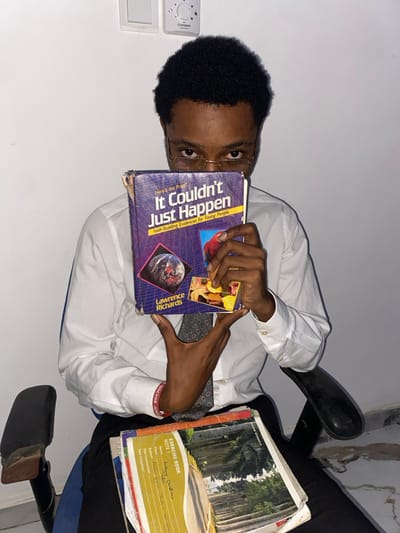 A young man in a white shirt and tie holds a book titled "It Couldn't Just Happen" by Lawrence Richards. The book's cover features a globe and colorful images. Additional exercise books are visible, one labeled "EXERCISE BOOK NOTE 1." The man wears glasses and a wristband with text on it. He sits in a chair with a black back, positioned against a white wall with a visible switch.