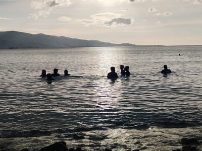 At Lamreh, Indonesia, a group of people are in the ocean. The scene is lit by the sun breaking through the clouds in the background. The background includes mountains in the distance. The water is wavy and dark, with light reflecting off the surface. There are rocks in the foreground. The people are scattered in the water, most are waist deep, with their upper bodies and heads visible.
