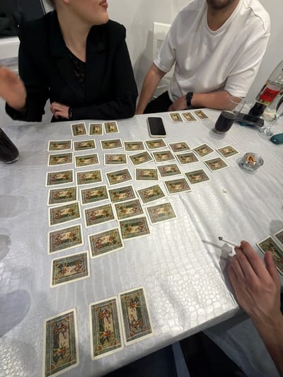 A high-angle shot depicts a group of people around a table covered in a white, textured tablecloth. Spread across the table are numerous playing cards, arranged in a pattern resembling a pyramid or a branching tree. To the left of the frame, a person in a black blazer is partially visible. Another person, wearing a white t-shirt, is positioned near the top left corner, next to a smartphone and a glass of dark liquid, possibly Coca-Cola. In the upper center, there's a small glass ashtray filled with cigarette butts. A hand holding a lit cigarette is visible on the upper right. A second person is seen on the upper right with denim attire. The location cannot be determined.