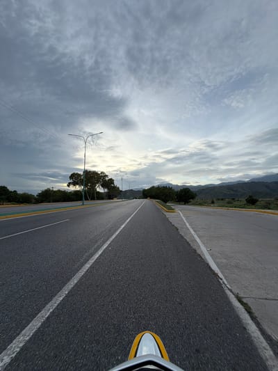 The image depicts an asphalt road with clear white lane markings, extending into the distance under a cloudy sky. On the left side of the road, a green buffer zone is visible, bordered by a yellow line. Beyond this, a line of trees and vegetation borders the road. In the background, rolling hills and mountains are visible, with the sky filled with textured clouds. Two lampposts are present along the roadside, and power lines stretch across the frame. The foreground shows the front of a yellow and silver vehicle, suggesting the image was taken from a moving vehicle. The scene appears to be a scenic stretch of highway, likely on the outskirts of a city, and given the context provided, it is identified as being in Maracay, Venezuela.