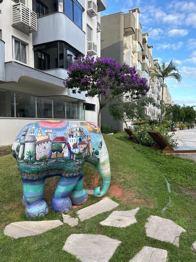 The image shows a colorfully painted elephant statue in a garden setting in Florianópolis, Brazil. The statue is predominantly painted with a townscape scene on its body and has teal and purple stripes on its legs and trunk. It stands on green grass, with a path of large stone slabs nearby. A building with a modern architectural design is visible in the background, featuring gray walls and large windows. A tree with purple flowers grows near the building. A taller building is visible further in the background. The sky above is blue with scattered white clouds.