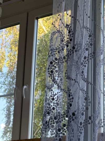 The image shows a close-up of a window with a sheer, embroidered curtain hanging in front of it. The curtain is white with a delicate pattern of dark blue or gray floral and circular motifs. Through the window, trees with bright yellow and green leaves are visible, suggesting it is autumn. The sky appears blue. The window frame is white, and there are two white handles on the window. The scene is indoors, with the curtain obscuring a direct view of the outside environment but allowing light and the colors of the foliage to filter through. The location is stated as Яромінскі сельскі Савет, Belarus. There are no people or animals visible in the image. The focus is on the texture and detail of the curtain and the interplay of light and shadow.